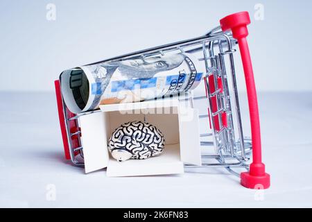 Capovolto carrello della spesa con cervello umano all'interno di una scatola di spedizione aperta e contanti isolato sul grigio. Concetto creativo di dipendenza dello shopping. Foto Stock