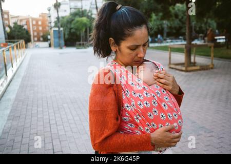 Giovane mamma che indossa il babbywearing in città. bambino in porta-confezione durante il trasporto. Maternità moderna e naturale all'aperto Foto Stock