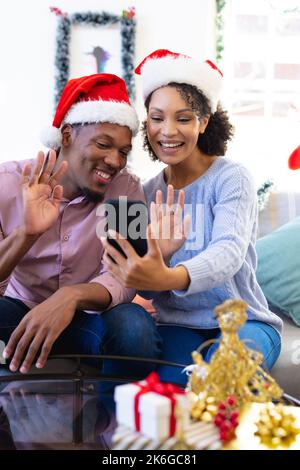 Coppia afro-americana felice che indossa cappelli di babbo natale, usando lo smatphone Foto Stock