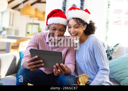 Coppia afro-americana felice che indossa cappelli di babbo natale, usando il tablet Foto Stock