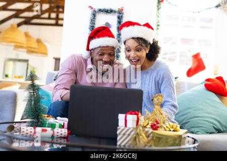 Felice coppia afro-americana che indossa cappelli di babbo natale, utilizzando un computer portatile Foto Stock