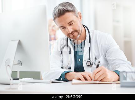 Compilazione di nuovi record. un medico maturo scrivendo note mentre lavora su un computer in un ufficio medico. Foto Stock