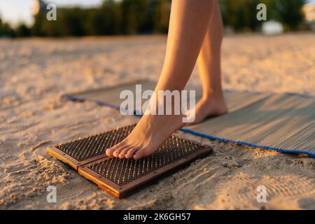 Piedi ravvicinati di una donna irriconoscibile che sale sul bordo del chiodo di Sadhu durante la pratica di meditazione di concentrazione sulla spiaggia sabbiosa del mare alla mattina di sole di estate Foto Stock