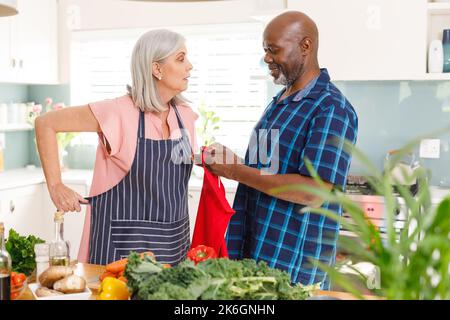 Felice coppia anziana diversa indossando grembiuli in cucina Foto Stock