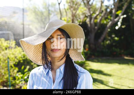 Ritratto di felice donna biraciale in cappello da sole in piedi in giardino soleggiato sorridente Foto Stock