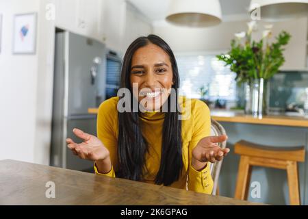 Donna felice biracial fare videochiamata sorridendo e gesturing alla macchina fotografica in cucina Foto Stock