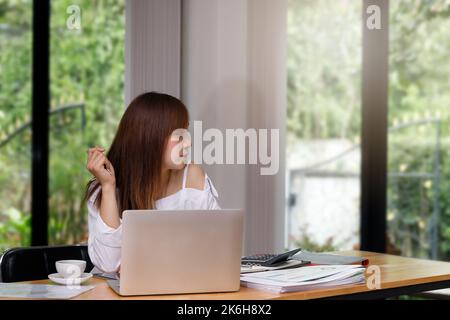 Giovane donna asiatica in abbigliamento casual elegante con notebook che guarda fuori dalla finestra in ufficio a casa. Foto Stock