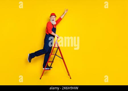 Foto a tutta lunghezza dell'età funky positiva uomo indossare tuta uniforme rosso elmetto scala da passeggio spazio vuoto isolato giallo sfondo Foto Stock