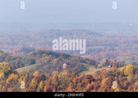 Case di villaggio sulla collina circondata da una lussureggiante foresta di colori autunnali, colline lontane svaniscono nella nebbia mattutina Foto Stock