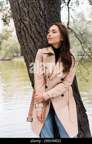 felice giovane donna in beige trench cappotto appoggiato sul tronco d'albero in parco, immagine stock Foto Stock