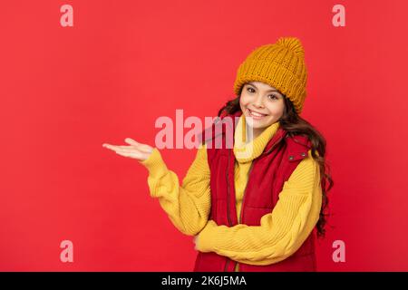 ragazza felice teen in cappello di inverno che presenta il prodotto su sfondo rosso, spazio di copia, pubblicità Foto Stock