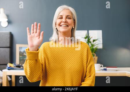 Donna anziana caucasica felice che fa videochiamata a casa sorridendo e ondeggiando alla macchina fotografica Foto Stock