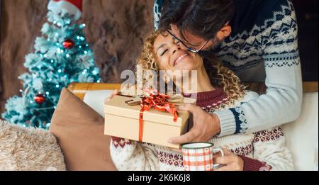 Felicità e amore persone in regalo di natale scambio attività di svago a casa. L'uomo che dà il regalo di sorpresa ad una donna overjoyed ed eccitata che siede o Foto Stock