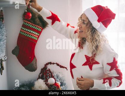 Donna sorridere e godere decorare appartamento con decorazioni natalizie. Le giovani donne indossano il cappello di babbo natale e si divertono durante il mese di dicembre ho Foto Stock