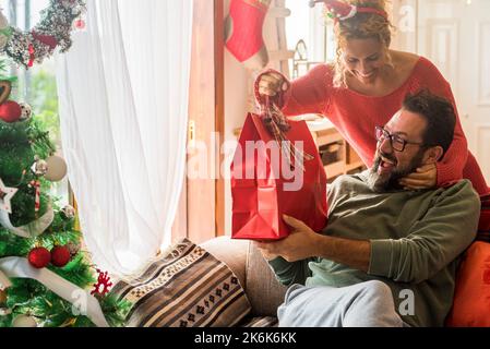 Persone a casa durante la celebrazione di natale. Felice donna sorridere e dare una sorpresa regalo a suo marito con gioia e divertimento. Coppia eccitata godere di Natale Foto Stock