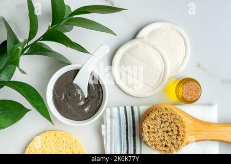 Vista dall'alto degli accessori benessere - spugne riutilizzabili, olio naturale, spazzola per massaggiare a secco, maschera di argilla e asciugamano di cotone su sfondo di legno Foto Stock