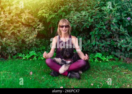 Donna blonde di mezza età con occhiali seduti sul prato con il suo cane pastore catalano che esegue meditazione ed esercizi di yoga. Sport con animali. Foto Stock