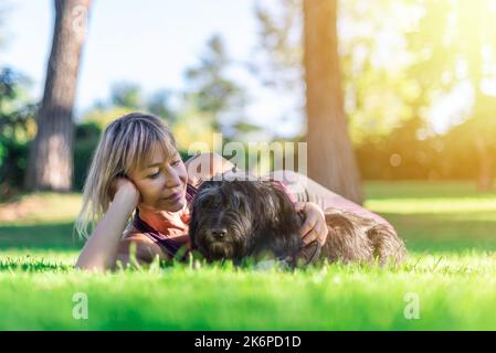 Donna bionda di mezza età sdraiata sull'erba e contemplando con amore il suo cane pastore catalano. Complicità e amore per gli animali. Gli animali domestici portano tranquillità Foto Stock