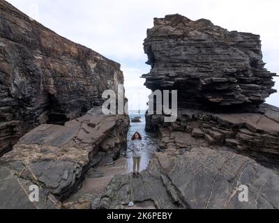 Donna con impermeabile che si stacca da sola in un paesaggio di scogliere e rocce in bassa marea con il mare sullo sfondo. Foto Stock
