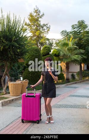 Una donna adulta felice in un vestito corto con una grande valigia rosa brillante è in piedi accanto alle palme, un viaggio di vacanza. Foto Stock