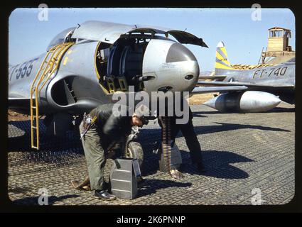 Vista frontale di tre quarti del naso di una Lockheed F-80C Shooting Star (s/n 47-555) recante le marcature del 80th Fighter Bomber Squadron (FBS), in qualche parte in Corea. Due dei pannelli sul naso sono aperti, e due crewmen a terra caricano le pistole con munizioni calibro .50. Una scala è montata contro il fianco dell'aereo, che conduce al pozzetto. Sullo sfondo, una parte della fusoliera posteriore e della coda di un'altra F-80C Shooting Star è visibile (s/n 49-740). Dietro questo velivolo appaiono dei sacchi di sabbia e una torre. Foto Stock