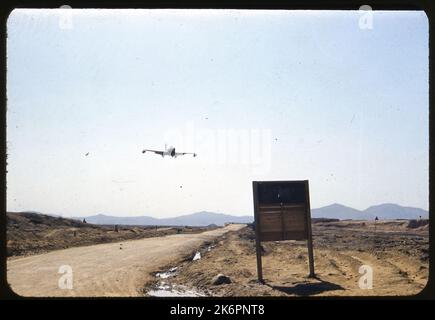 Vista frontale a metà destra di una Lockheed F-80 Shooting Star che si avvicina alla pista, da qualche parte in Corea. L'aeromobile è dotato di serbatoi laterali. In primo piano è un segno di legno, che si trova in una zona fangosa accanto alla pista. Sullo sfondo, un uomo si siede su una piccola collina che guarda l'aereo. Foto Stock