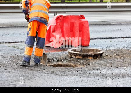 Un lavoratore della strada in tute arancioni libera la bocca di una botola fognaria sulla carreggiata con una pala per la sua successiva riparazione. Spazio di copia. Foto Stock