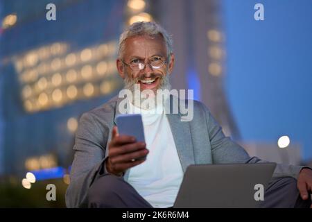Buon vecchio uomo d'affari seduto fuori ufficio nel parco cittadino utilizzando il telefono di notte. Foto Stock