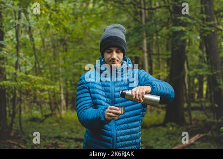 Escursionista su un lungo sentiero prende un riposo e versa il tè caldo da un thermos in una tazza per riscaldare e rifornire vitamine e liquidi. Escursioni in alta montagna. Foto Stock