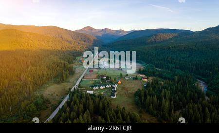 Vista aerea di mattina di nebbia luminosa sopra le piccole case rurali tra cime scure con alberi di foresta di montagna all'alba di autunno. Bellissimo scenario di selvaggio Foto Stock