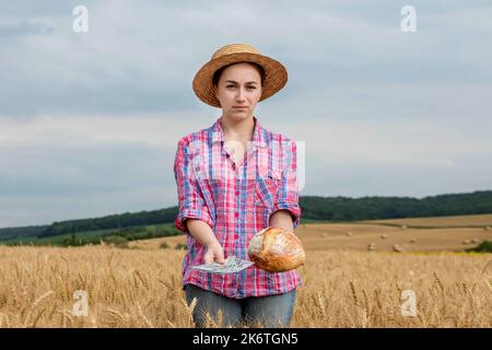 Giovane agricoltore che tiene dollari e pane in campo di grano dorato. Profitto dall'agricoltura durante la stagione di raccolta in estate. Foto Stock