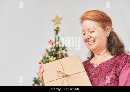 Felice donna latina matura sorridendo mentre tiene il regalo di Natale che ha appena ricevuto, a casa. La gioia delle vacanze. Foto Stock