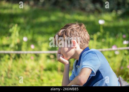 Rovesciato piccolo ragazzo caucasico si sente male triste annoiato pensieroso seduto da solo in un parco, bambino pensare nascondere problema Foto Stock