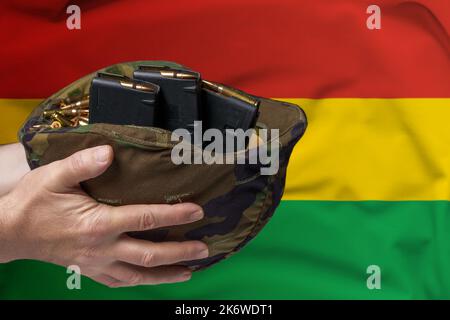 Un casco militare con cartucce e riviste per un fucile nelle mani di un uomo sullo sfondo della bandiera della Bolivia. Il concetto di Foto Stock