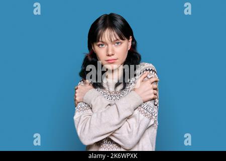 Giovane donna congelata in maglione con mani stringate gestendo freddo, su sfondo blu Foto Stock