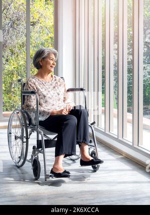 Donna anziana asiatica felice in abito casual si siede in sedia a rotelle, sorriso e guardando fuori dalla finestra di vetro con vista verde naturale, stile verticale. Posizioni Foto Stock