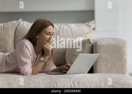 Bella donna sdraiata sul divano utilizzando un computer portatile a casa accogliente Foto Stock
