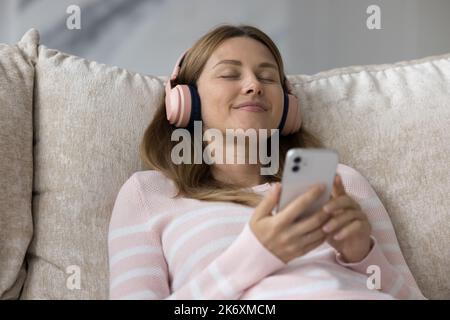 Donna pacifica in cuffie tiene il cellulare rilassante sdraiato sul divano Foto Stock