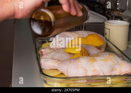 Vassoio da forno con tre cosce di pollo su un letto di patate, cipolla e spicchi di limone, e una mano con un olio può versare olio sulle cosce. Foto Stock