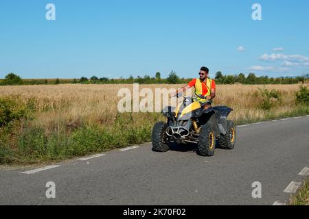 Giovane uomo che guida veicoli ATV su strada asfaltata campagna tra campi di erba in Transilvania, Romania il 17 luglio 2022 Foto Stock