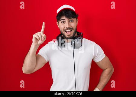 Uomo ispanico con barba che indossa cappello da giocatore e cuffie che puntano il dito verso l'alto con un'idea di successo. uscito e felice. numero uno. Foto Stock