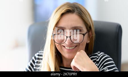 Donna d'affari felice e sofisticata di mezza età seduta sulla sedia da ufficio, sorridente e guardando la macchina fotografica Foto Stock