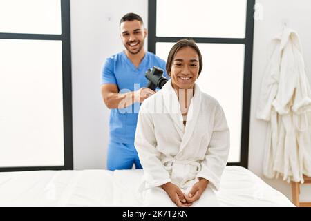 Latino uomo e donna che indossano la fisioterapia uniforme massaggiante collo con pistola a percussione al centro di bellezza Foto Stock