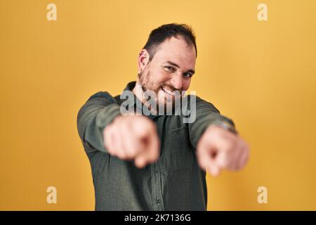 Più misura uomo ispanico con barba in piedi su sfondo giallo puntando a voi e la macchina fotografica con le dita, sorridendo positivo e allegro Foto Stock