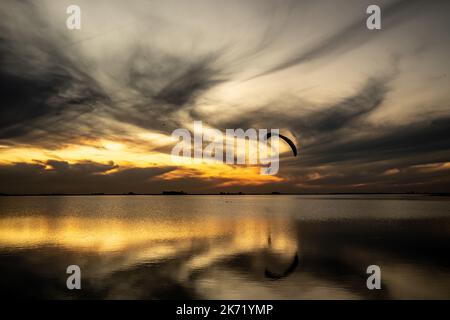 Paesaggio lagunare durante il tramonto. Un parapendio motorizzato si avvicina mostrando la sua silhouette contro la luce. Foto Stock