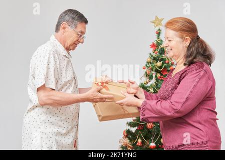 Coppia latina anziana sorridente, trascorrendo le vacanze a casa, scambiando regali di Natale vicino a un albero decorato. La gioia di celebrare insieme. Foto Stock