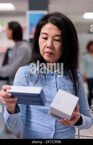 Donna asiatica guardando i pacchetti supplementi in farmacia negozio, esaminando scatole di vitamine e bottiglie di farmaci sugli scaffali. Cliente farmacia che acquista prodotti medici e forniture. Foto Stock