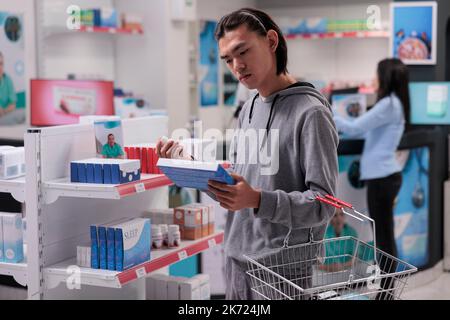 Cliente asiatico che esamina pacchetto farmaci per curare la malattia, guardando integratori medici sugli scaffali. Cliente farmacia che acquista prodotti farmaceutici per la salute, farmaci e pillole. Foto Stock