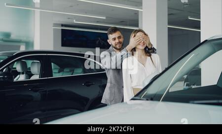 Ragazzo allegro bearded sta facendo sorpresa per la sua moglie che chiude gli occhi con le sue mani e che la conduce a bella nuova auto è motore showroom, donna felice è abbracciare e baciarlo. Foto Stock