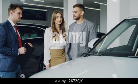 Il venditore amichevole dell'automobile in vestito sta comunicando con il giovane uomo sicuro che gli dice circa il nuovo modello del veicolo mentre la donna graziosa sta levandosi in piedi vicino al suo marito che tiene il suo braccio e sorridente. Foto Stock